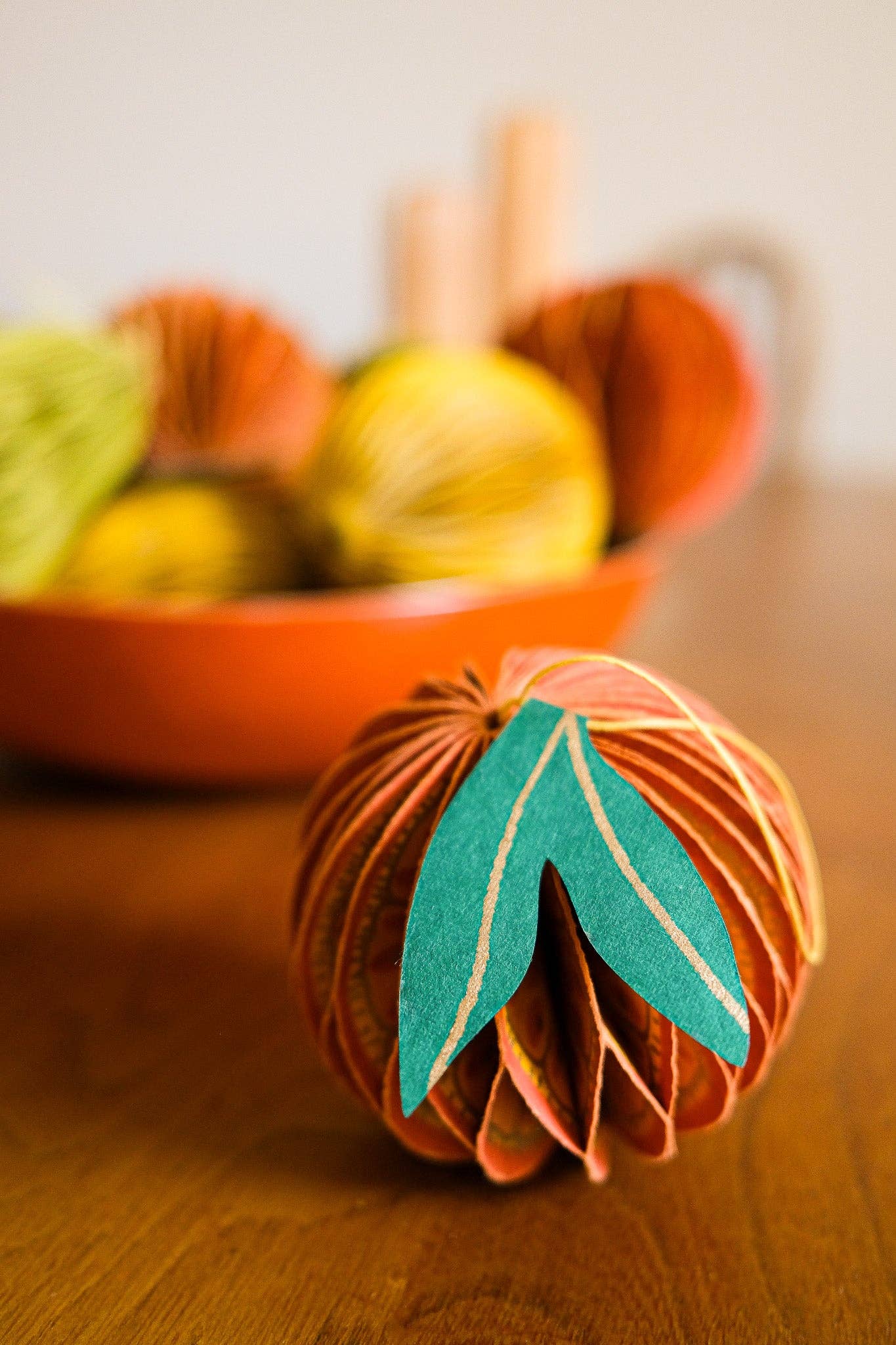 Decorative paper pumpkin with green leaf on a wooden surface, blurred yarn balls in the background.