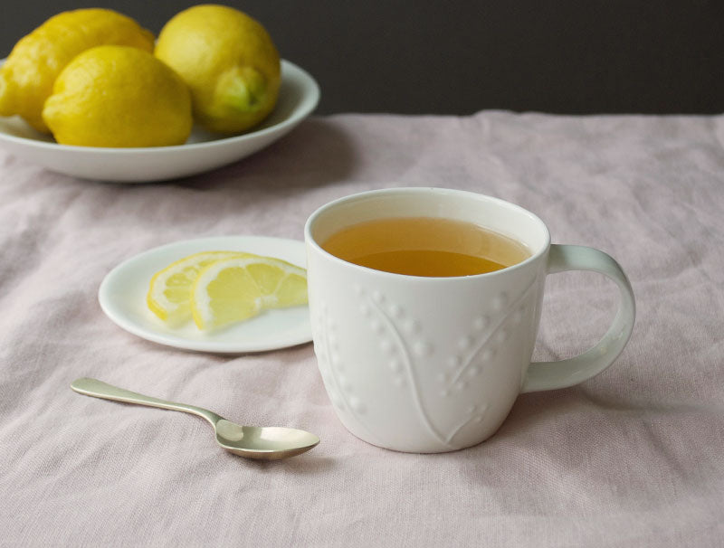 White mug with tea, lemon slices, and lemons on a tablecloth.