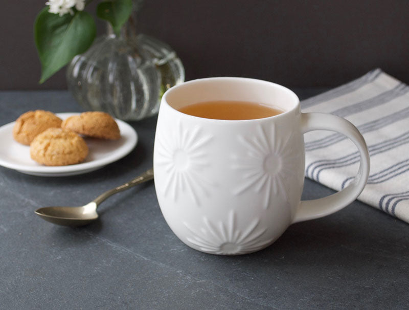 White embossed mug with tea on a dark surface with cookies and a vase in the background