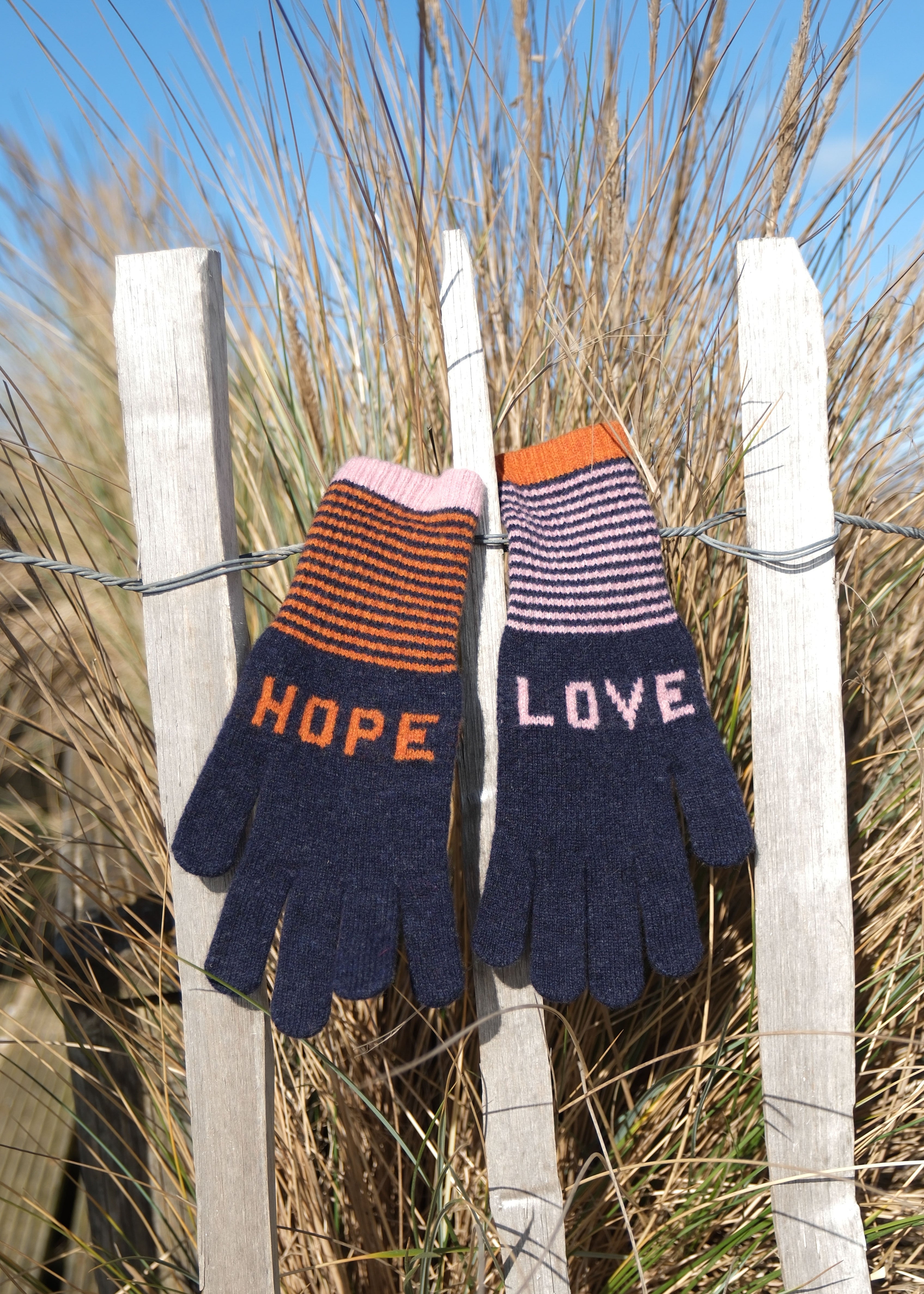 Pair of knitted gloves with 'HOPE' and 'LOVE' text hanging on a fence with grass in the background.