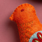 Close-up of an orange knitted or felted animal toy with embroidered details on a pink background