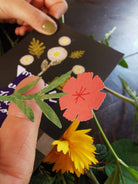Hand holding a black card with floral designs and a red flower cutout, surrounded by green leaves and a yellow flower.