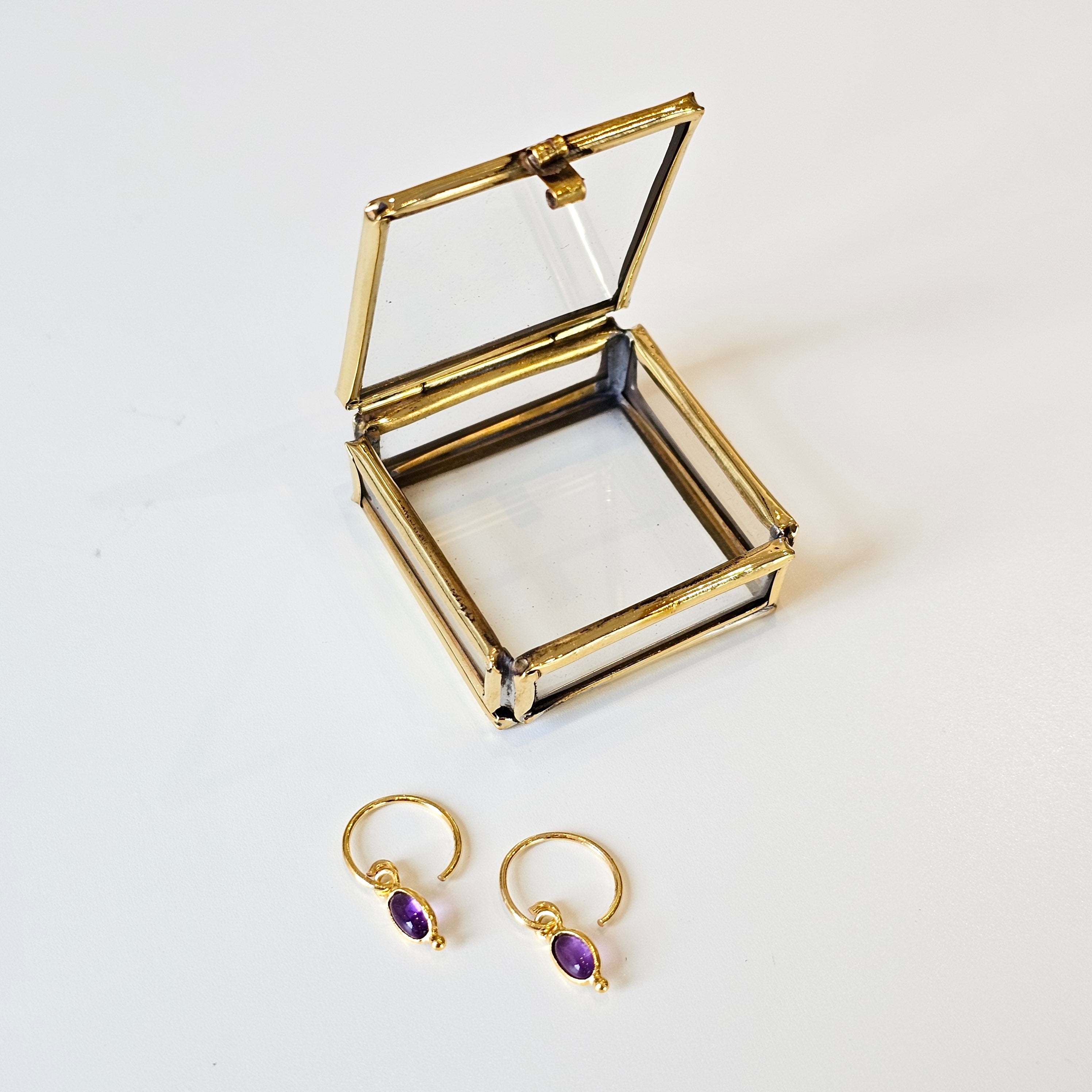 Gold earrings with amethyst stones next to an open gold jewellery box on a white background