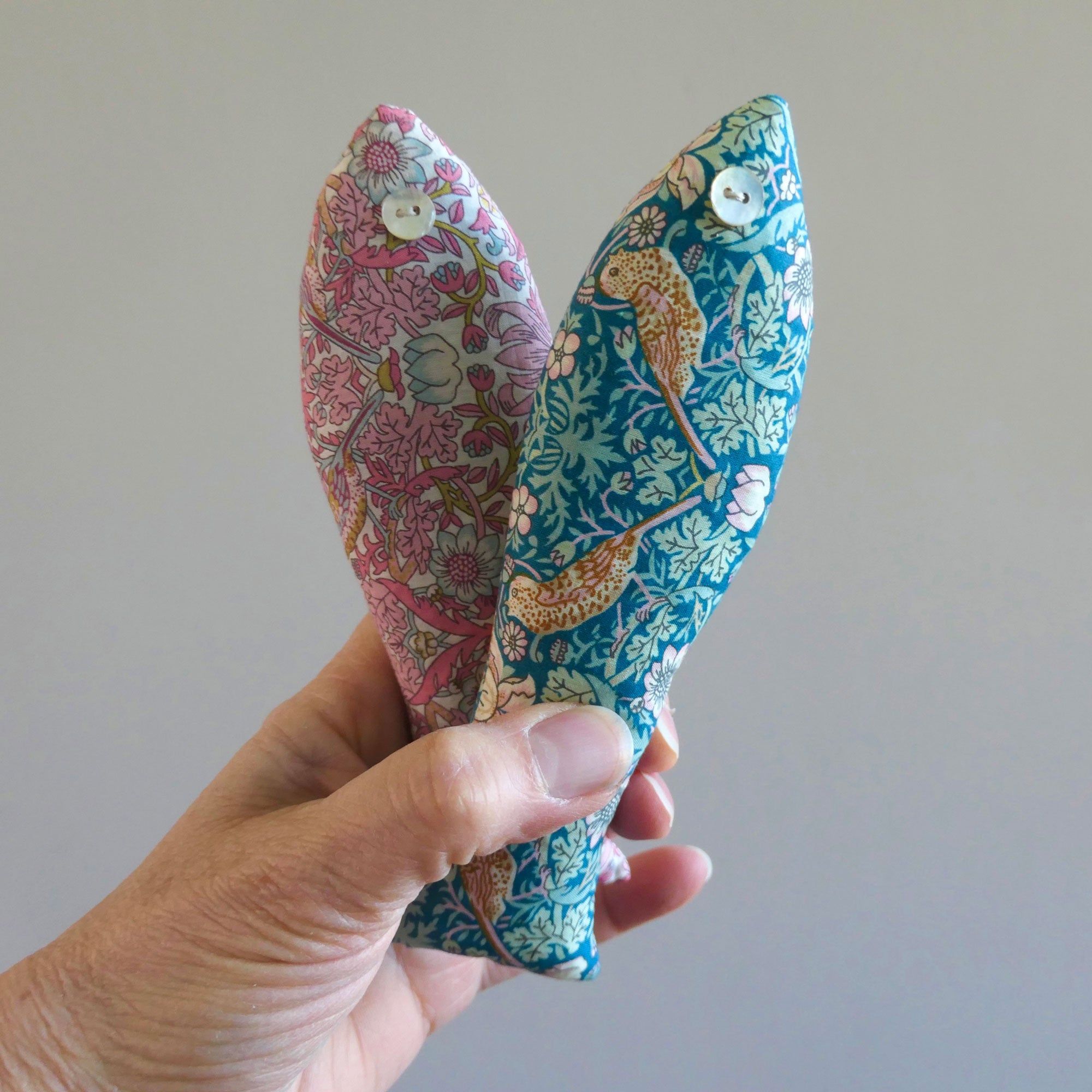 Hand holding two floral-patterned fish-shaped sachets in Strawberry Thief Liberty Fabric against a plain background