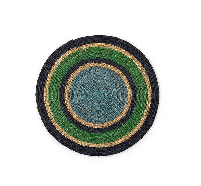A round woven placemat with concentric rings of black, green, teal-blue, and natural tan jute on a plain white background.