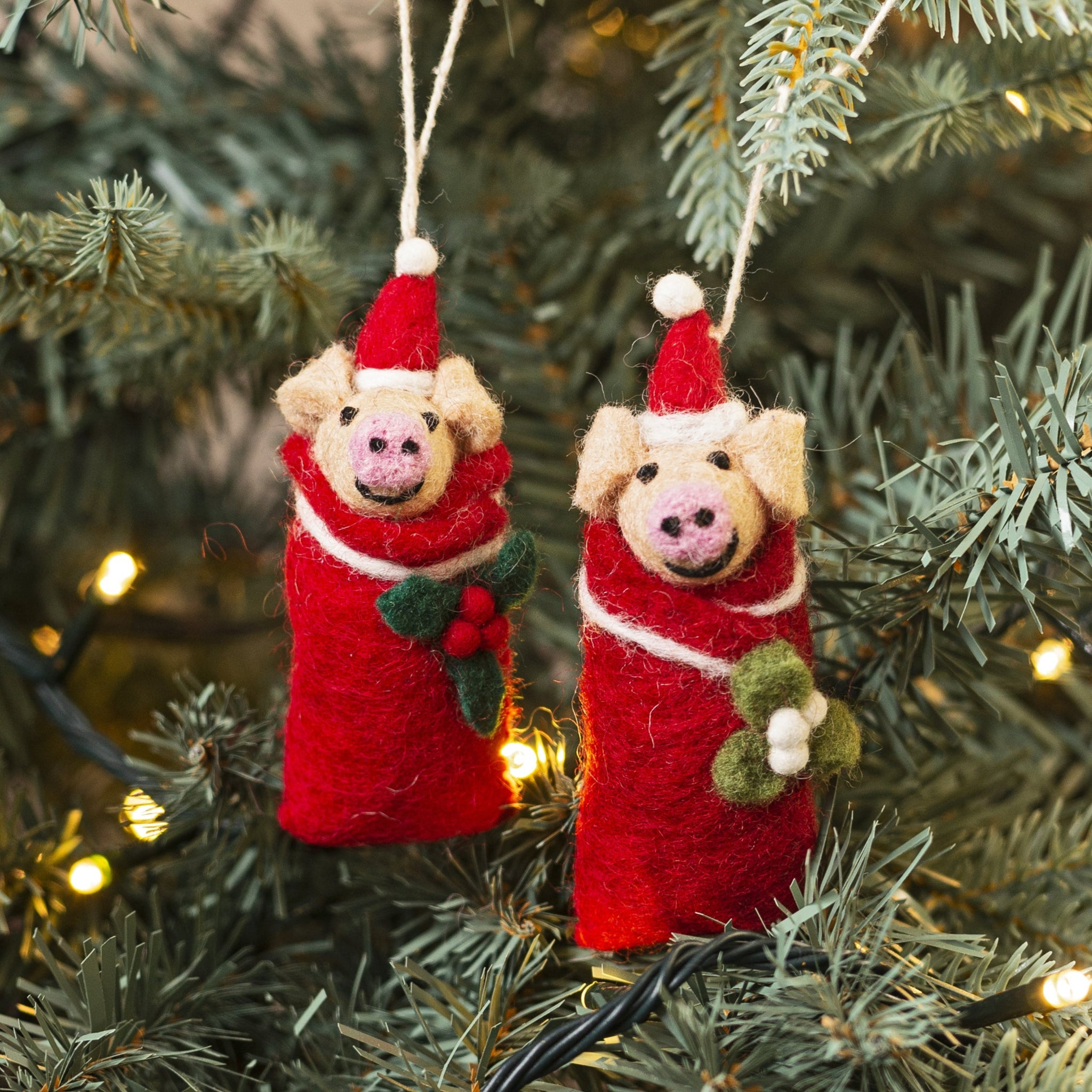 Two felted pig ornaments in Santa outfits hanging on a Christmas tree.