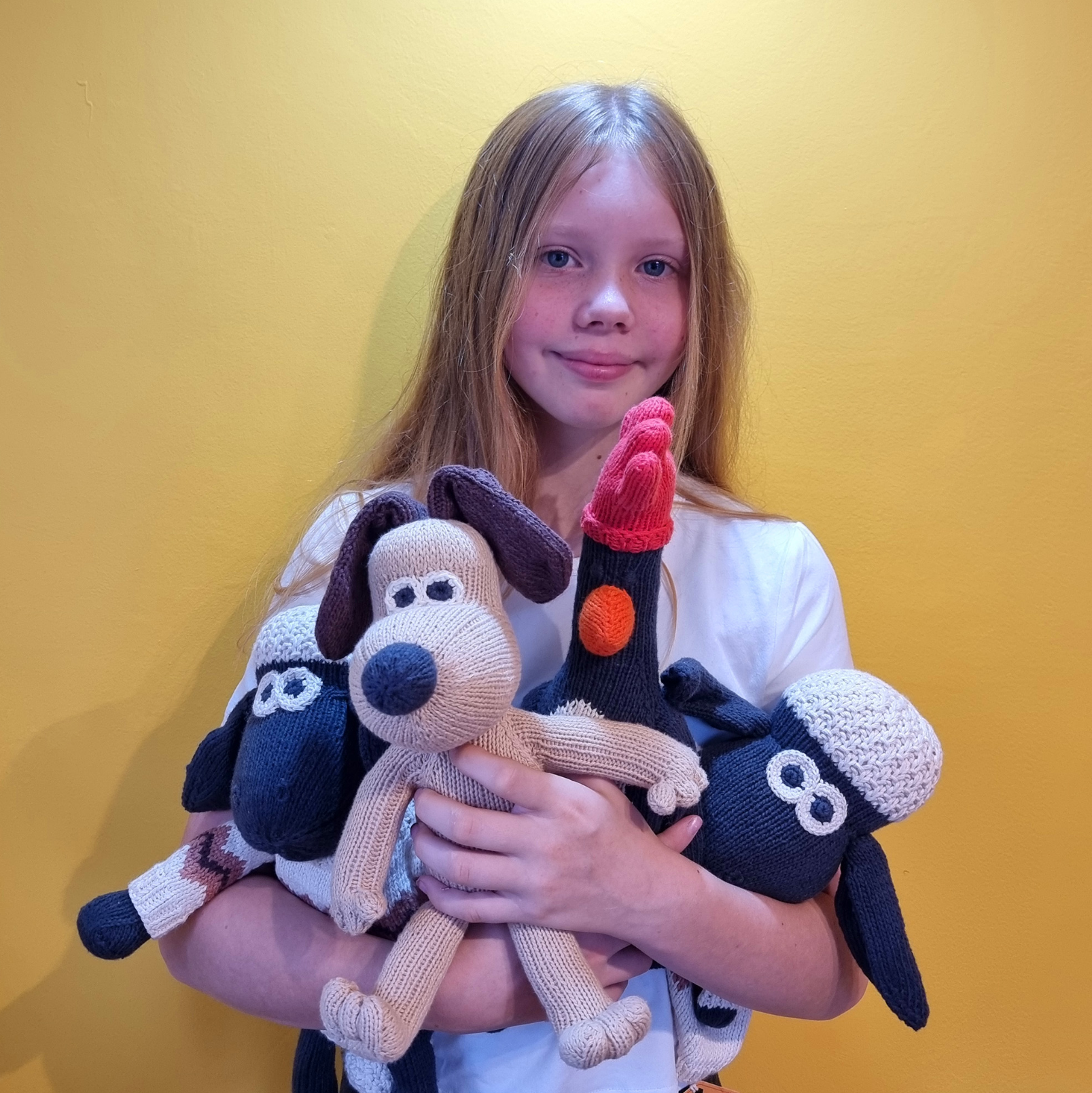 A young girl with long light brown hair holds three crocheted plush toys against a solid yellow background. The toys include a beige and black dog, a red and black bird, and a blue and white sheep.