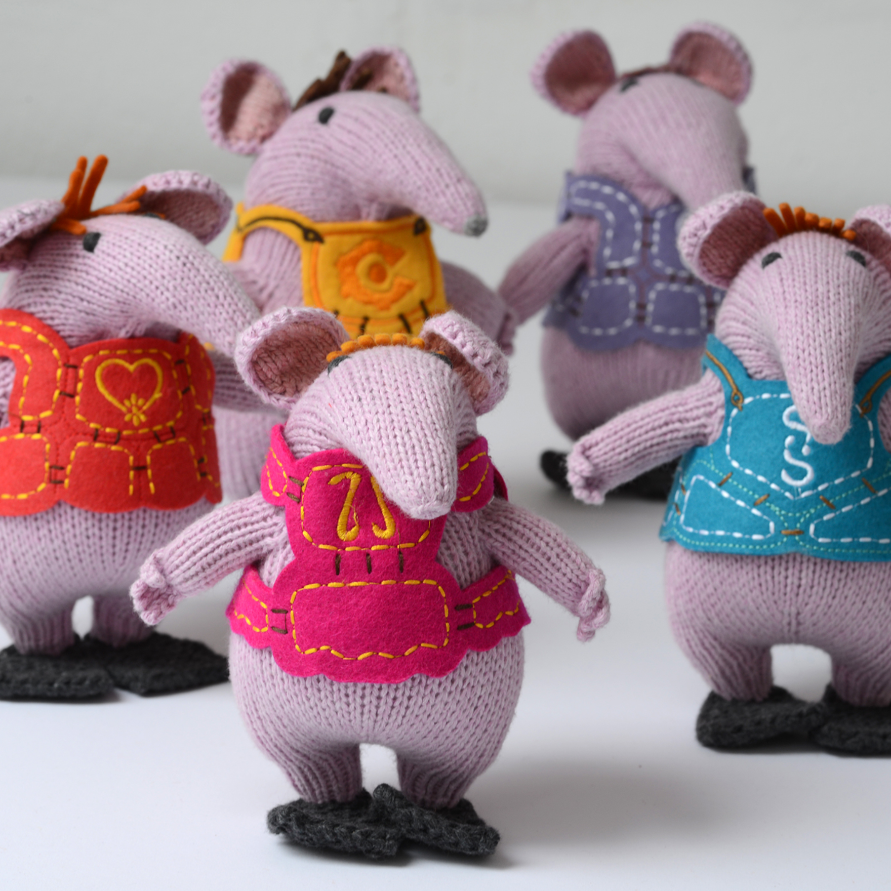 Five knitted mouse plush toys standing side by side, each wearing a different coloured vest. The vests are orange-red, yellow, pink, and blue, and the mice are made of light purple-pink knit fabric.