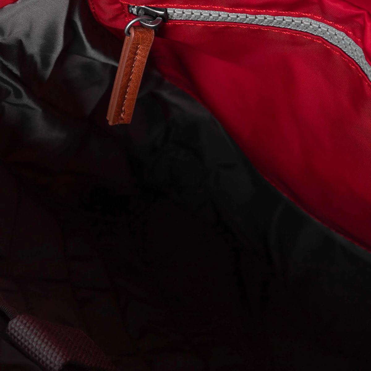 Close-up of the inside of a red bag with black lining, a grey zip with a brown leather pull tab on the inner pocket.