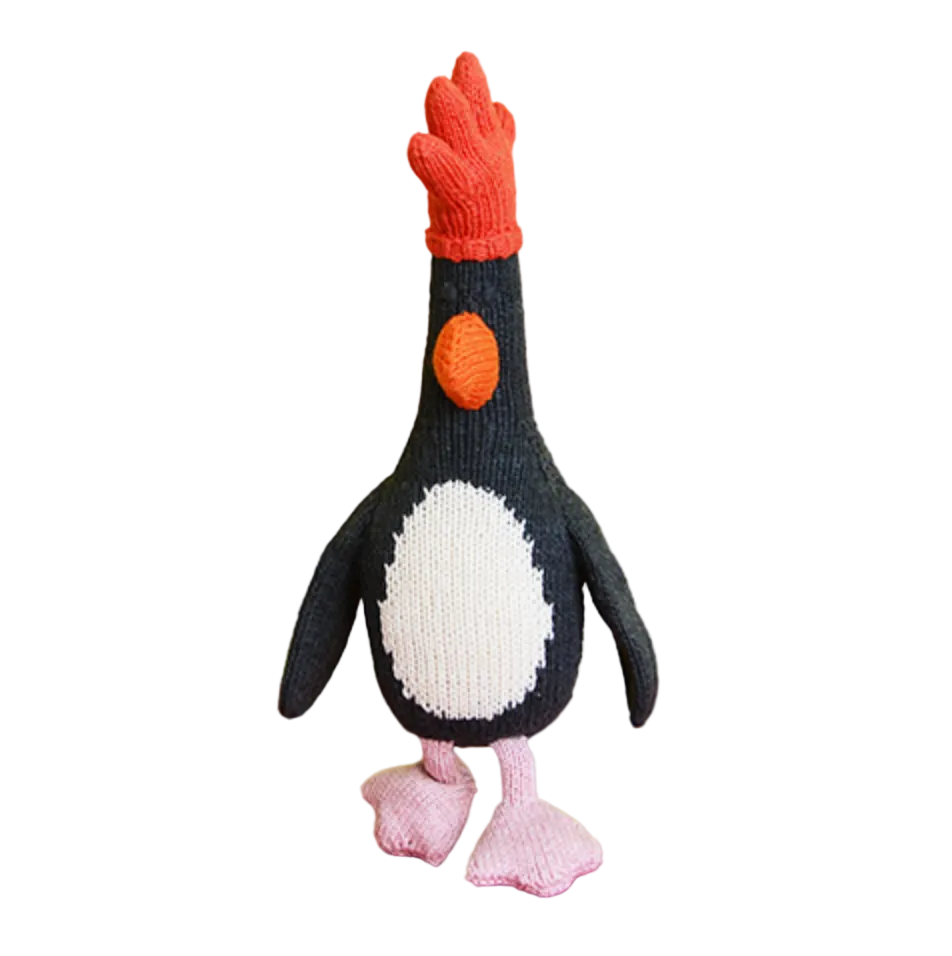 A crocheted penguin doll with a red comb, black and white body, and pink feet, displayed on a yellow and orange background.