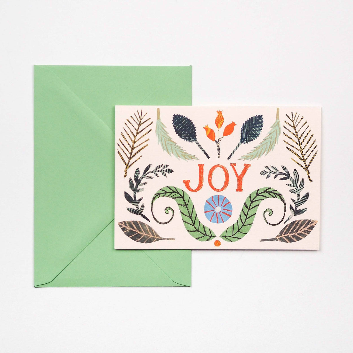 A greeting card with a botanical illustrations, placed in front of a green envelope on a light background. The card features the word “JOY” in orange-red lettering with decorative leaves and small floral motifs around it.