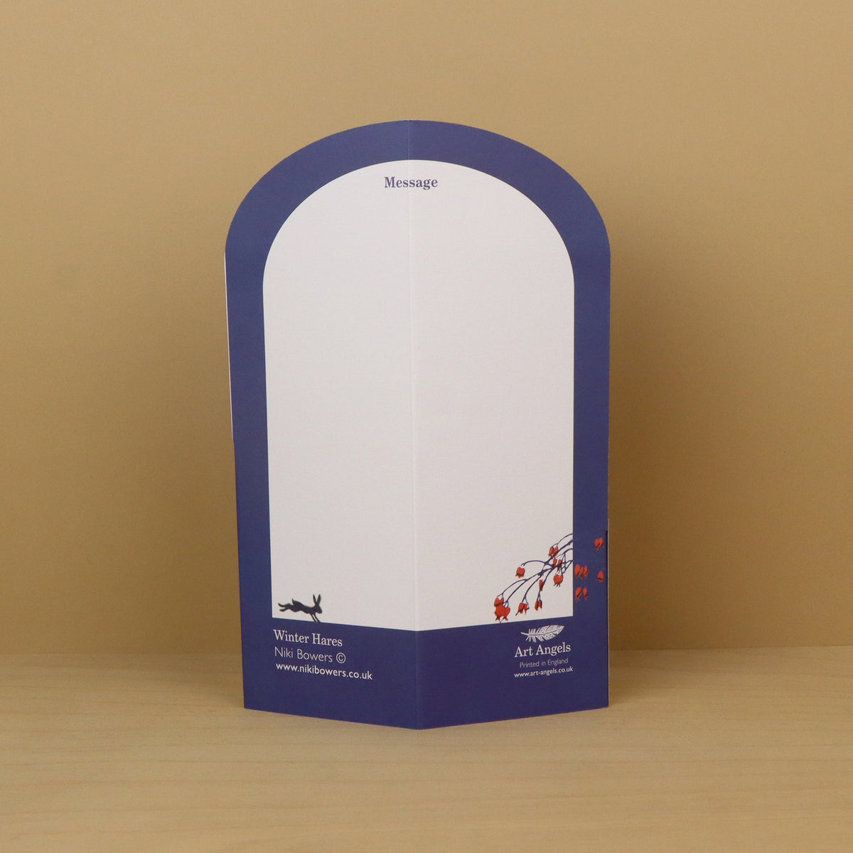 A freestanding arched paper card with a white writing area and a dark blue border, placed on a light wood surface against a tan background. The bottom section includes printed branding and contact details.