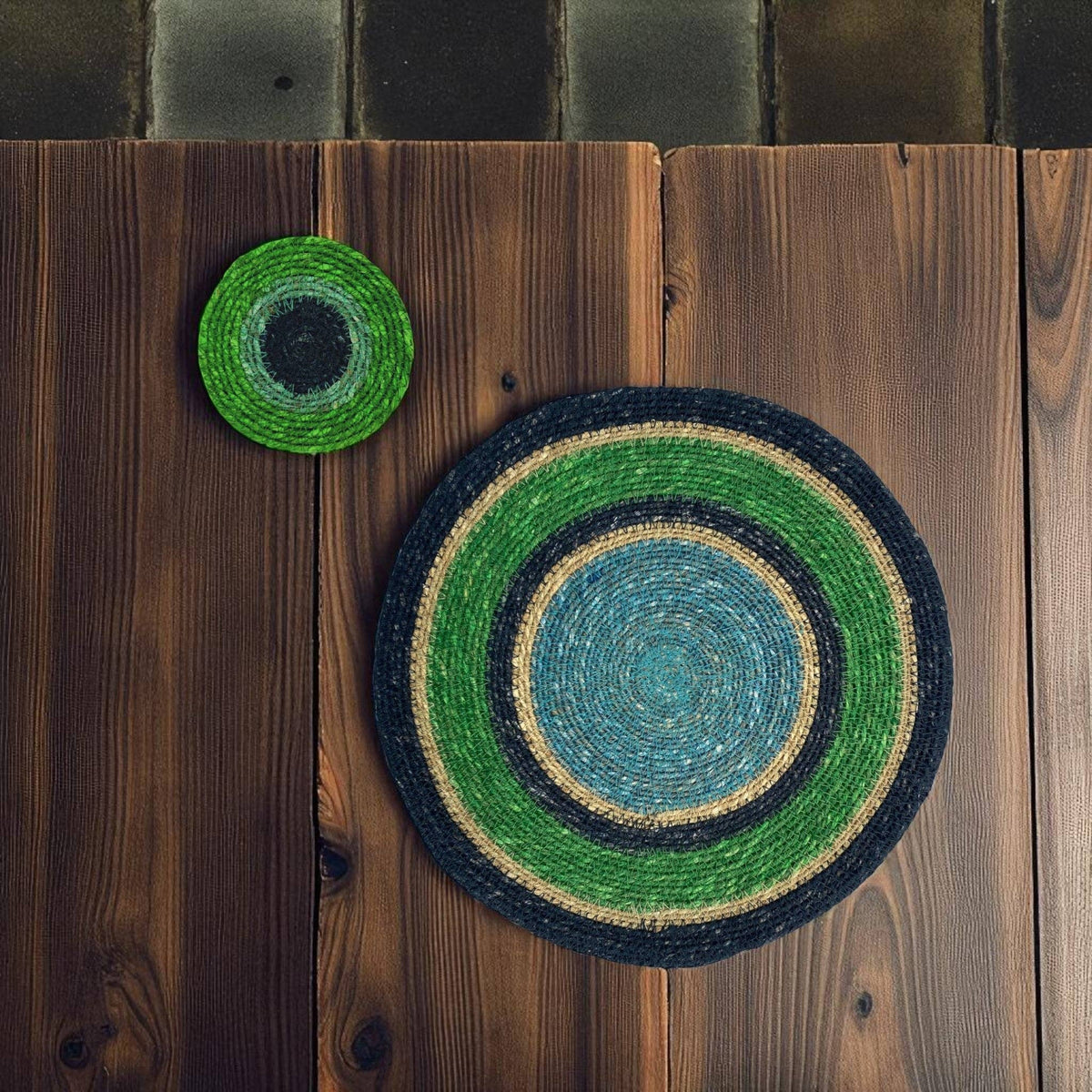 Two round woven placemats with concentric circular patterns in green, blue, black, and natural beige, displayed on a rustic dark brown wooden plank surface.