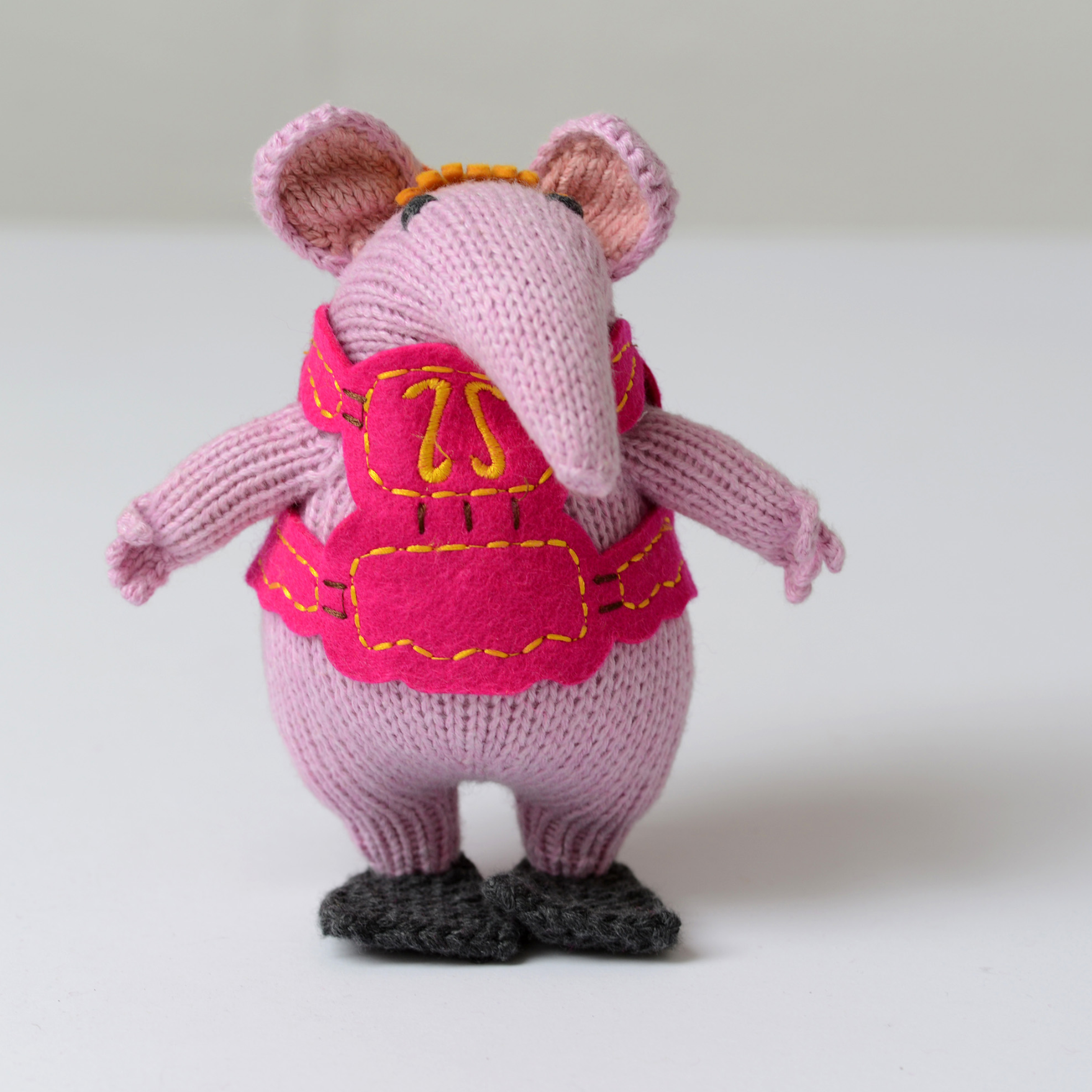 A knitted mouse figurine in light pink with a bright pink vest, yellow embroidered details, and dark gray shoes, shown against a plain light background.