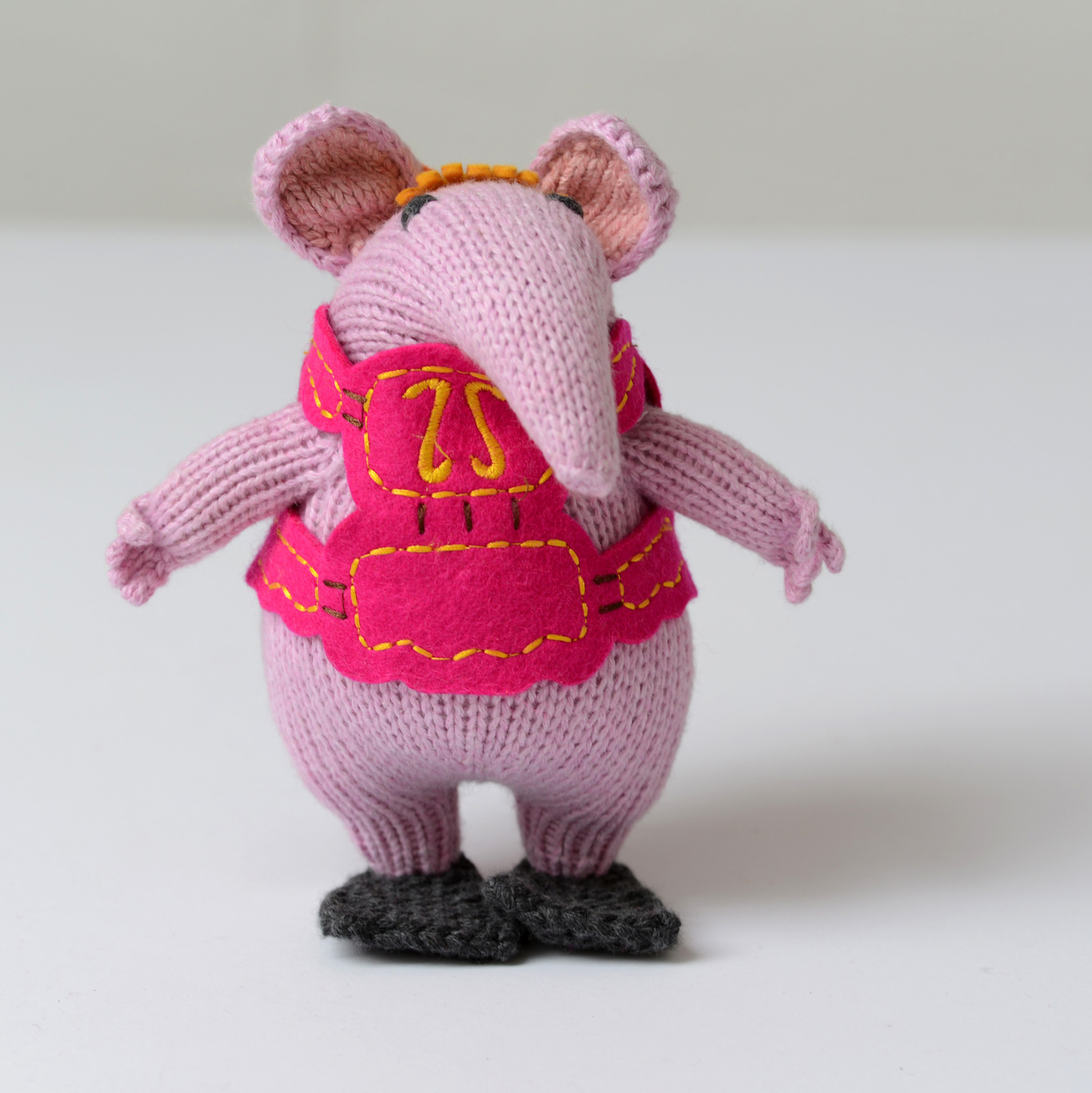 A knitted mouse figurine in light pink with a bright pink vest, yellow embroidered details, and dark gray shoes, shown against a plain light background.