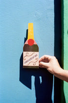 Hand holding a bottle-shaped 'Congratulations' card against a blue wall.