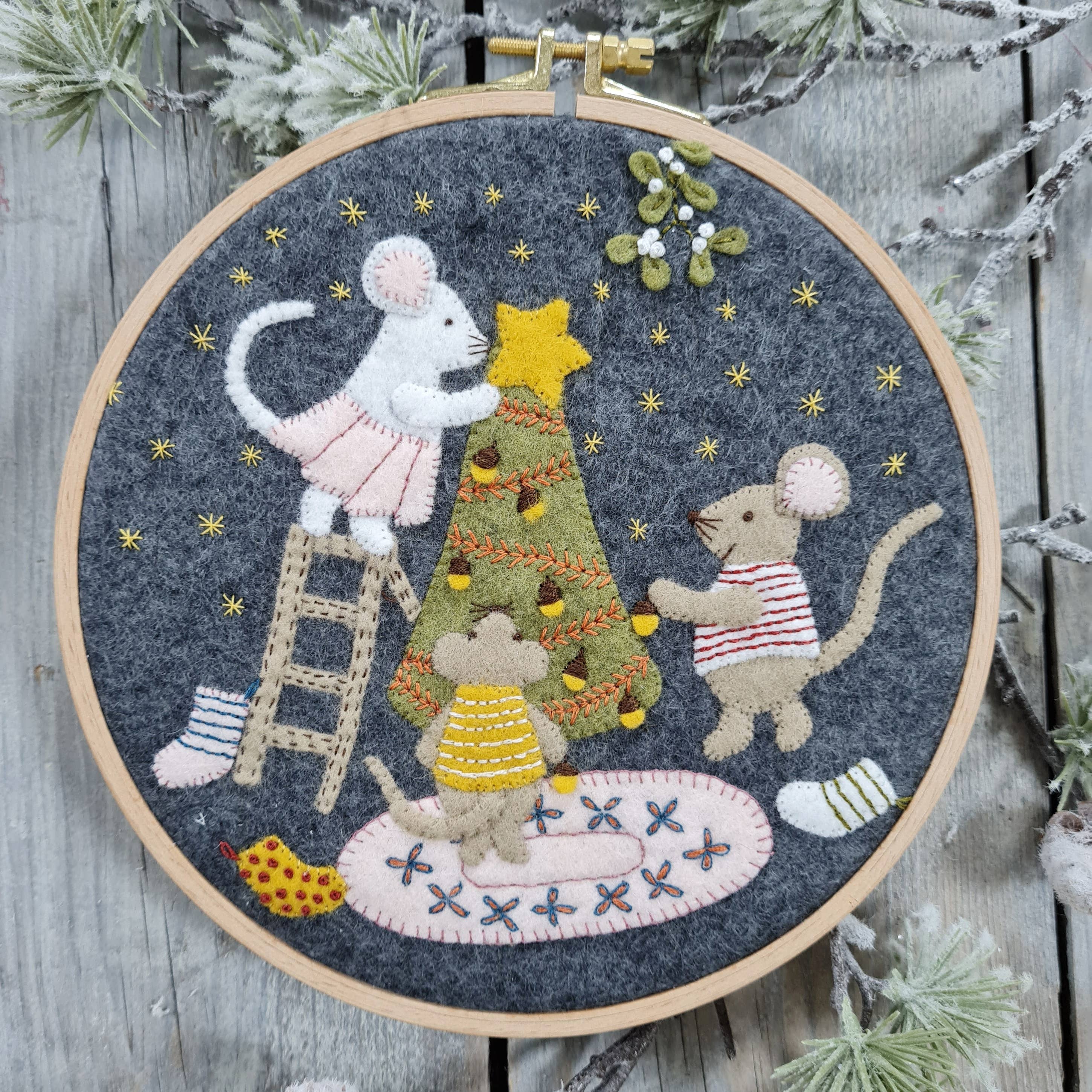 Embroidery hoop with festive scene of mice around a Christmas tree on a wooden background.
