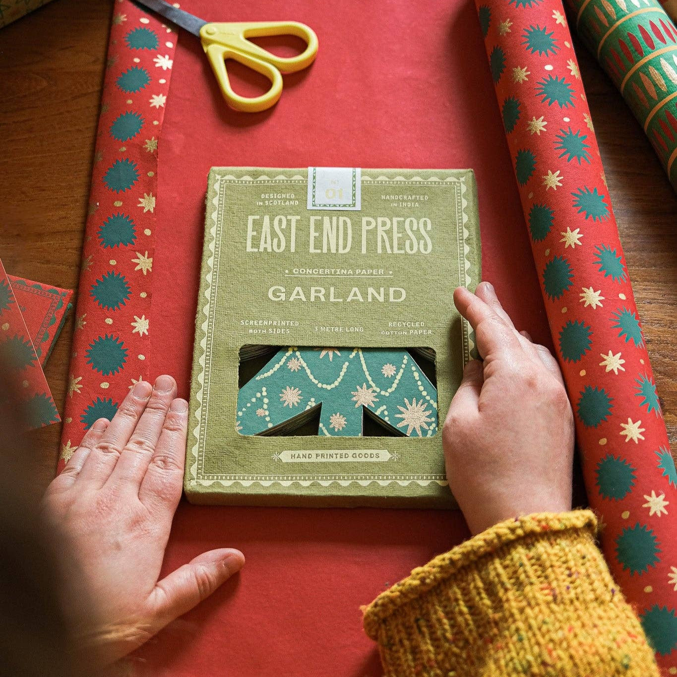 Person holding a package of 'East End Press Garland' on red wrapping paper with scissors.