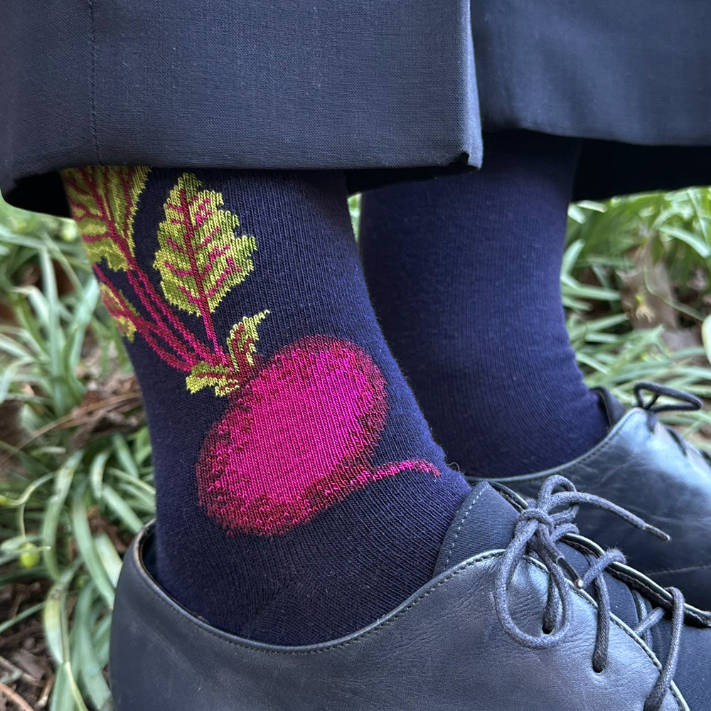 A close-up of a person's legs wearing navy blue socks with a large pink and green beechnut design, paired with dark gray dress shoes and dark trousers, set against a background of green plants.