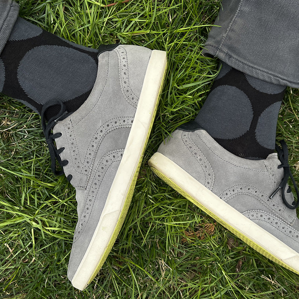 A top-down view of a person's feet wearing gray suede wingtip sneakers with off-white soles and black socks with gray polka dots, standing on green grass.