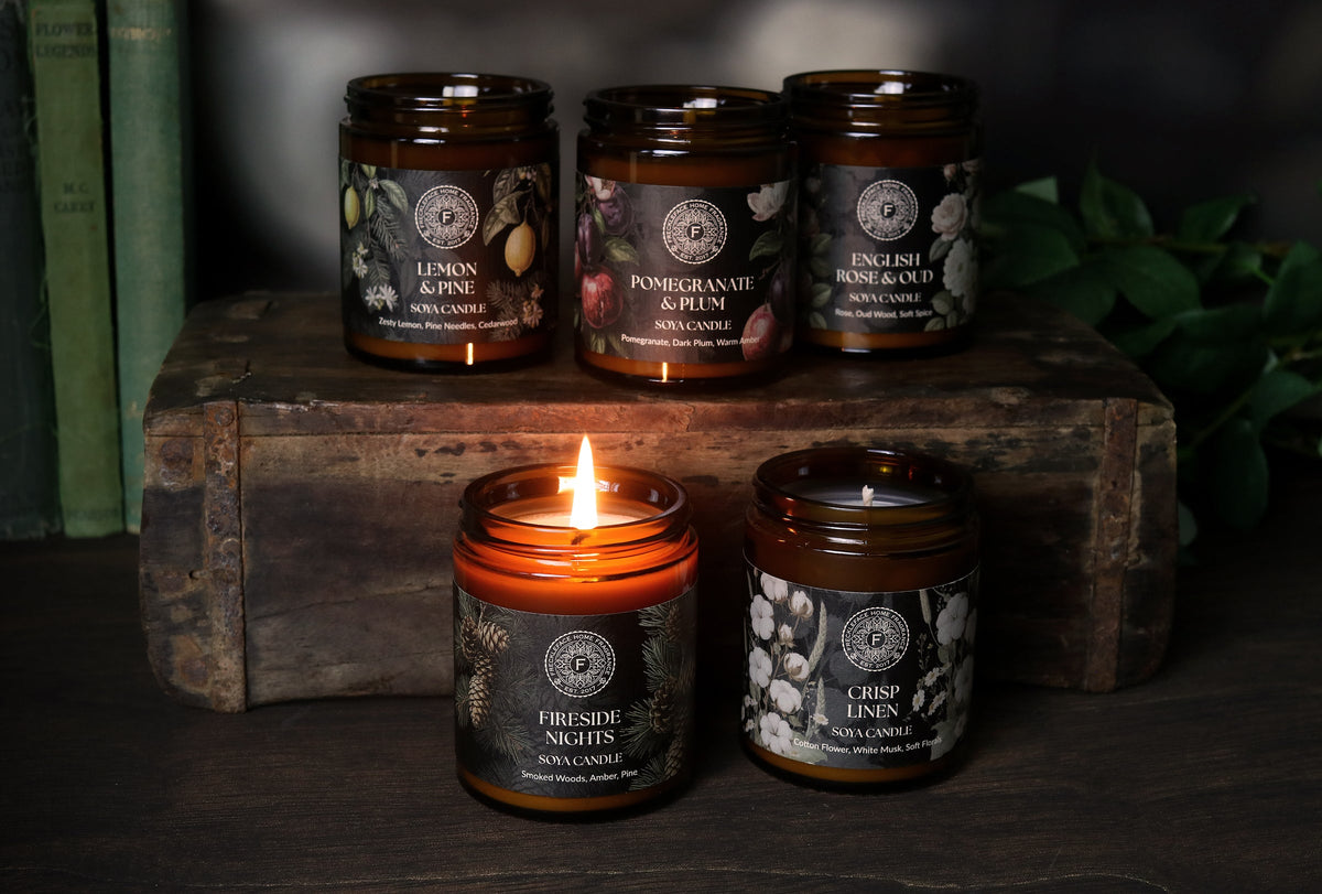 Four amber glass scented candles displayed on a rustic wooden crate, with one candle lit in the foreground. The labels feature botanical illustrations and scent names including Lemon & Pine, Pomegranate & Citrus, English Rose & Oakmoss, and Crisp Birch.
