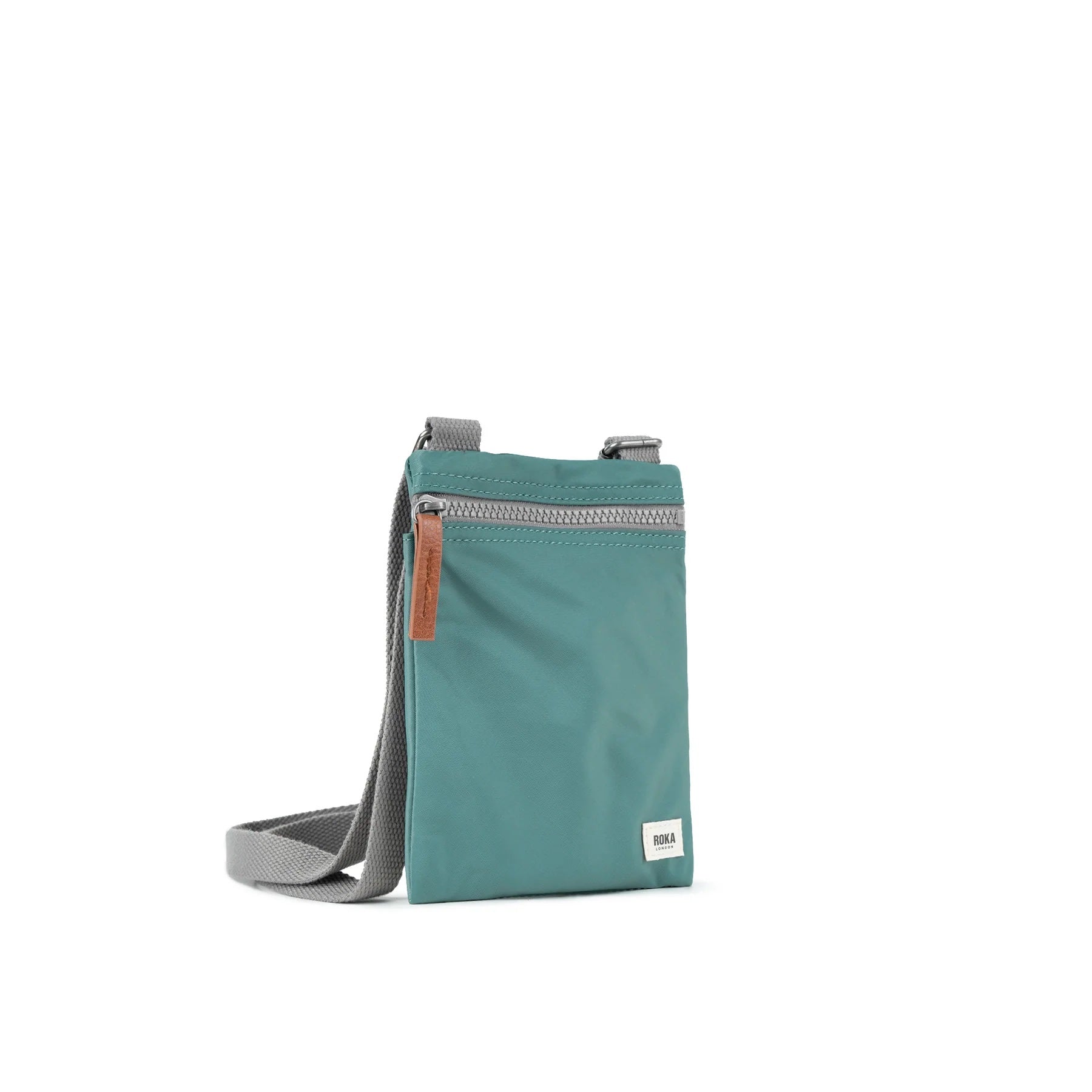 A photo of the front and side of a small rectangular sage green pocket bag. It has a grey zip horizontally at the top, grey straps, and a small ROKA logo in the bottom right corner. The zip pull is brown.