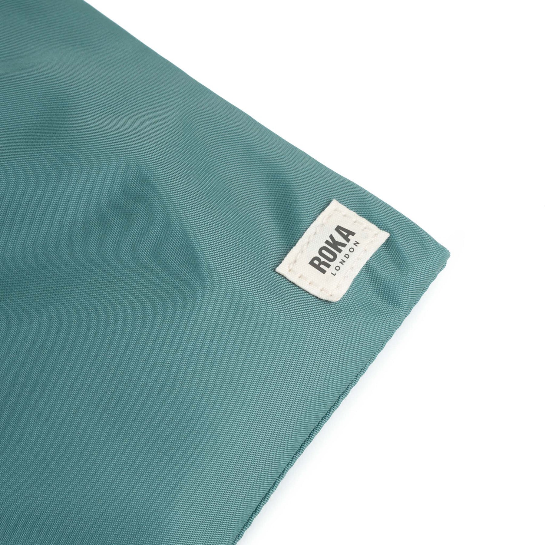 A close up photo of the bottom corner of a sage green bag, showing the ROKA London label.