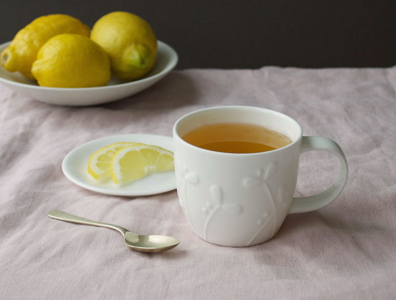 White mug with tea, lemon slices, and lemons on a tablecloth.
