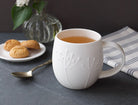 White mug with tea on a dark surface with cookies and a vase in the background