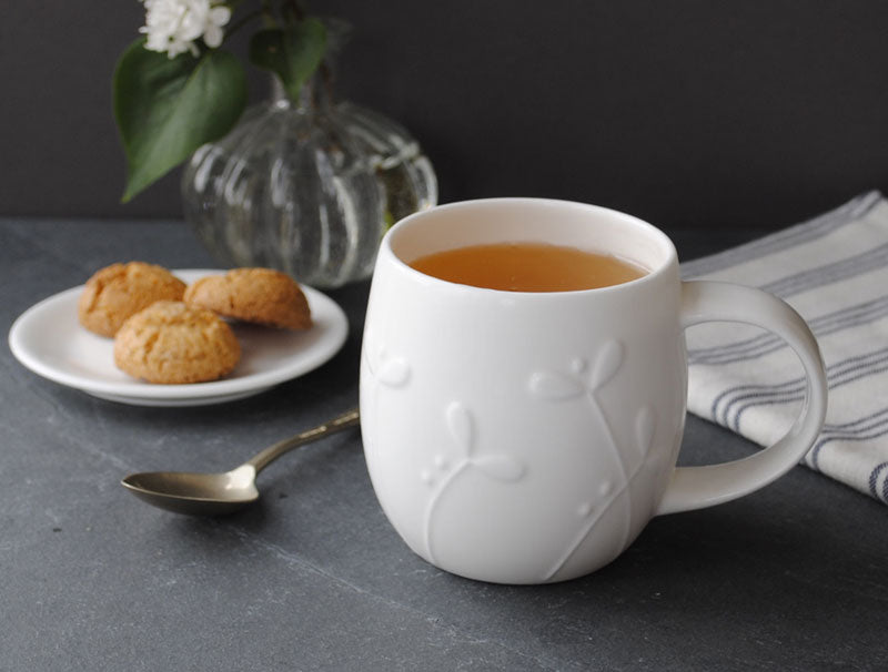 White mug with tea on a dark surface with cookies and a vase in the background