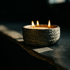 A rustic, textured ceramic candle holder with three lit wicks and glowing flames, set on a dark stone surface in dramatic, low-key lighting.