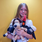 A young girl with long light brown hair holds three crocheted plush toys against a solid yellow background. The toys include a beige and black dog, a red and black bird, and a blue and white sheep.