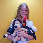 A young girl with long light brown hair holds three crocheted plush toys against a solid yellow background. The toys include a beige and black dog, a red and black bird, and a blue and white sheep.