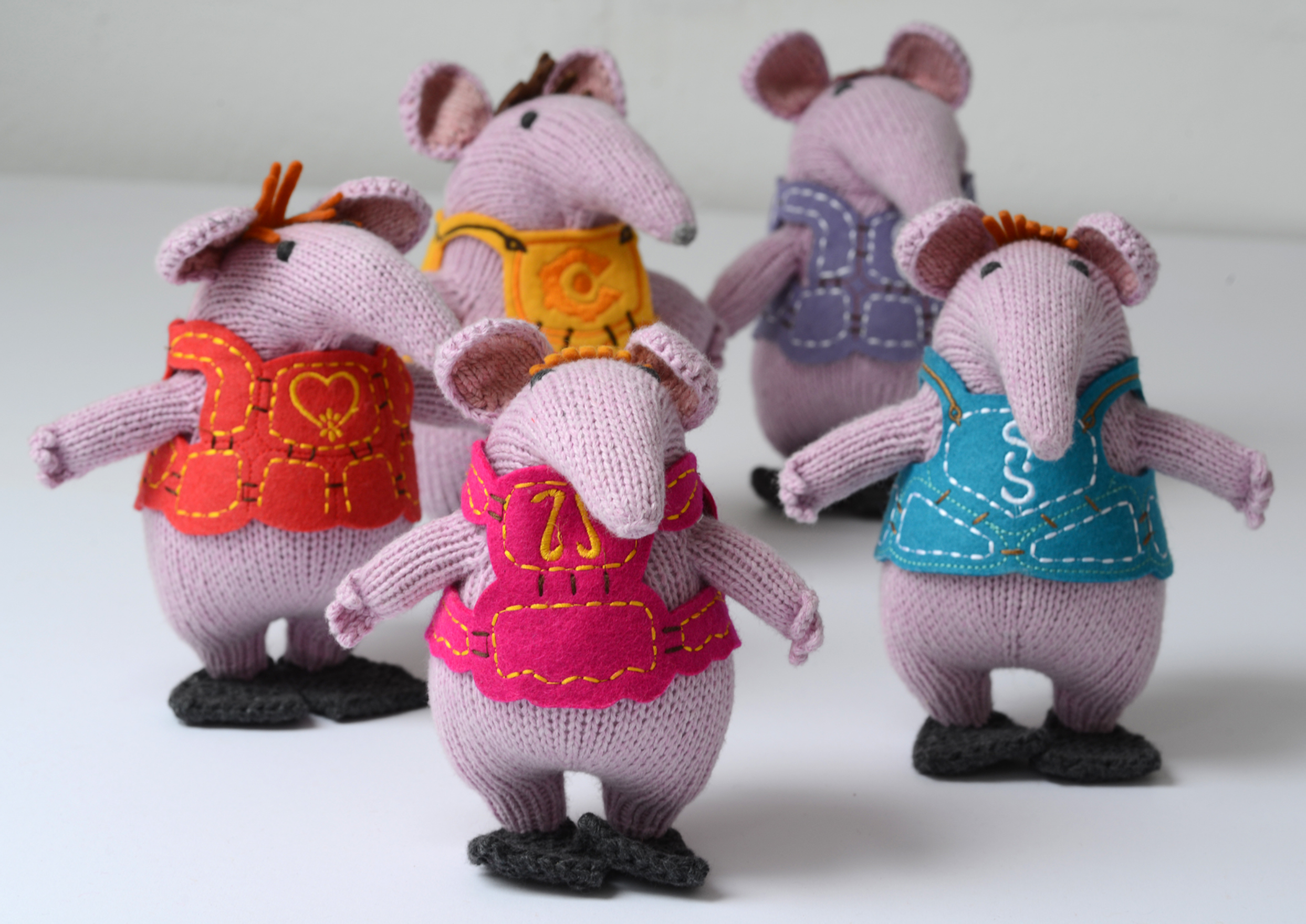 Five knitted mouse plush toys standing side by side, each wearing a different coloured vest. The vests are orange-red, yellow, pink, and blue, and the mice are made of light purple-pink knit fabric.