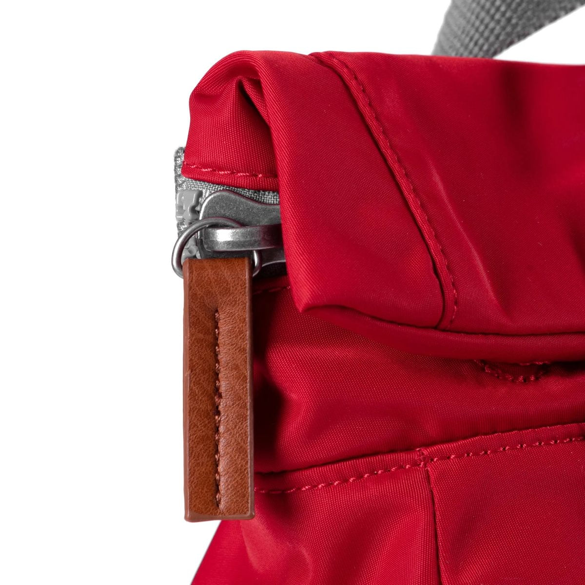 Close-up of a red bag, showing a grey zip with a brown leather pull, and a red closing flap on a white background