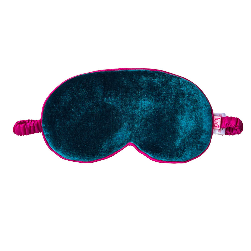 A plush velvet sleep mask in teal with a vibrant pink trim and matching elastic straps, shown against a plain white background.