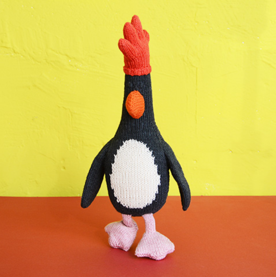 A crocheted penguin doll with a red comb, black and white body, and pink feet, displayed on a yellow and orange background.