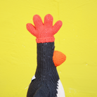 A knitted rooster hat or hat-like accessory with a bright red crown and orange beak, attached to a black knit body, shown against a yellow background.