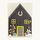 Decorative card with a fisherman’s house design featuring a horseshoe, windows, and plants on a beige background.
