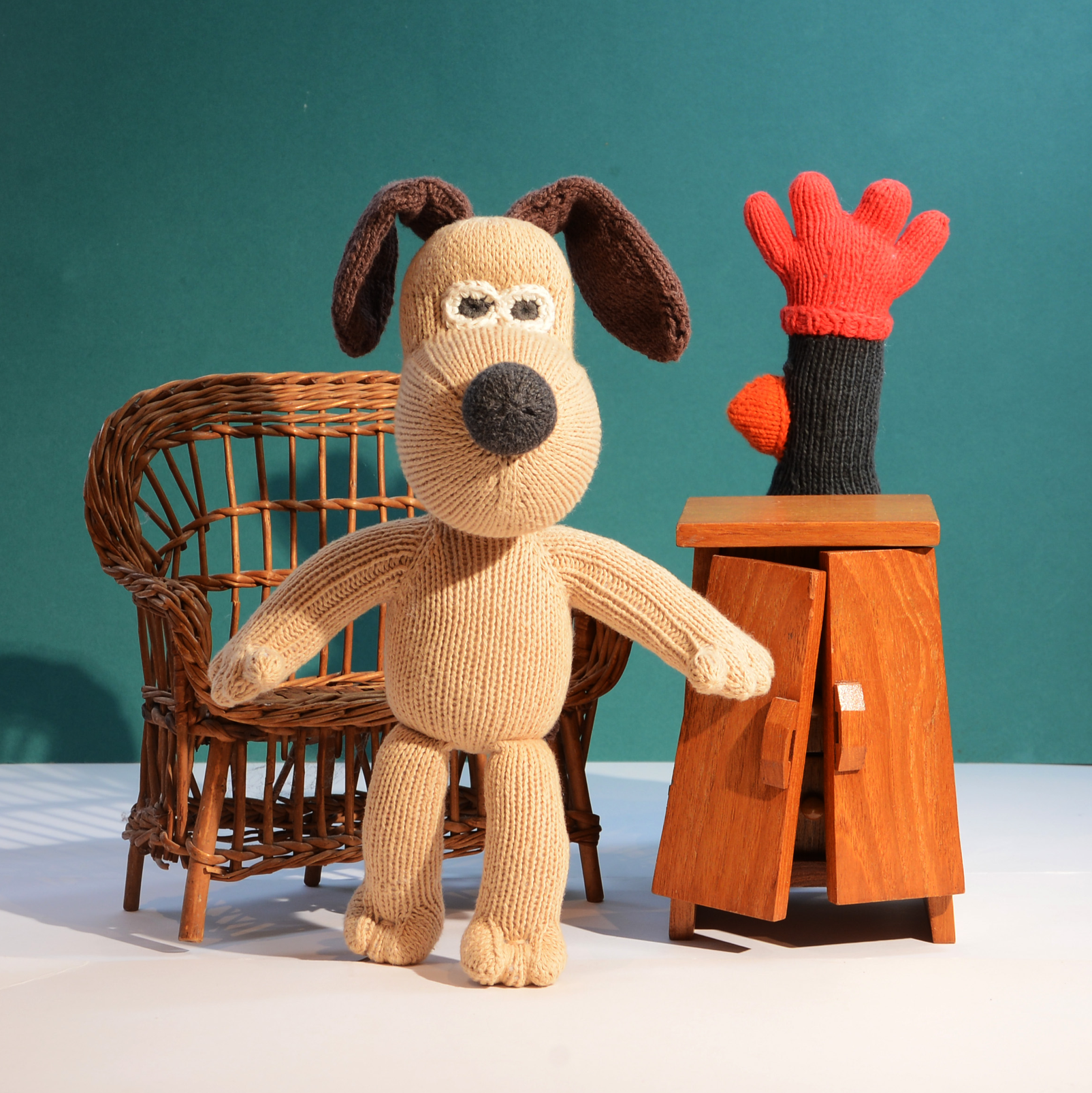 A knitted dog plush toy with floppy dark brown ears stands in front of a teal studio backdrop, next to a small wooden cabinet and a red-and-black glove prop.