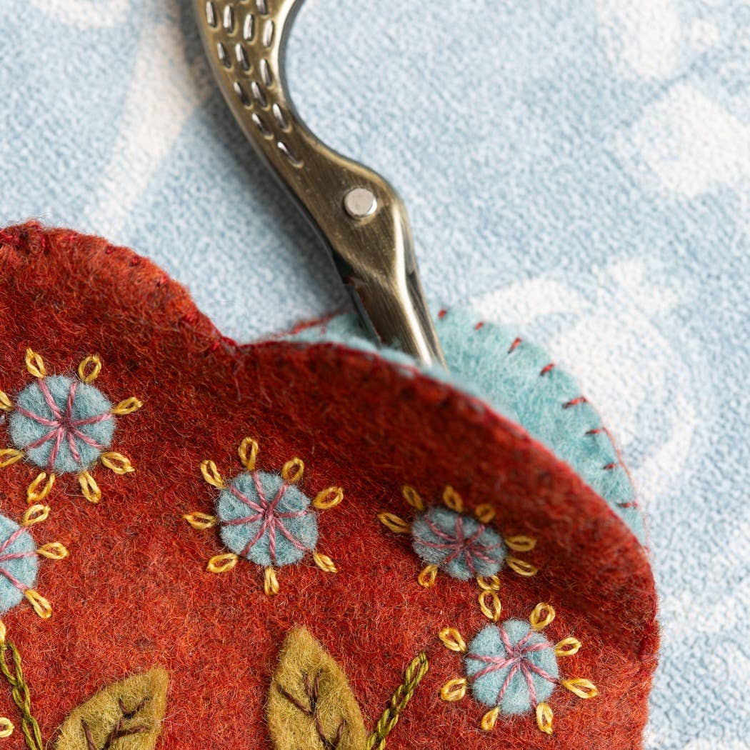 Detail of embroidered red heart scissors pouch, showing blue felt lining and red felt upper with colourful botanical embroidered details and scissor handles poking out of the opening. 
