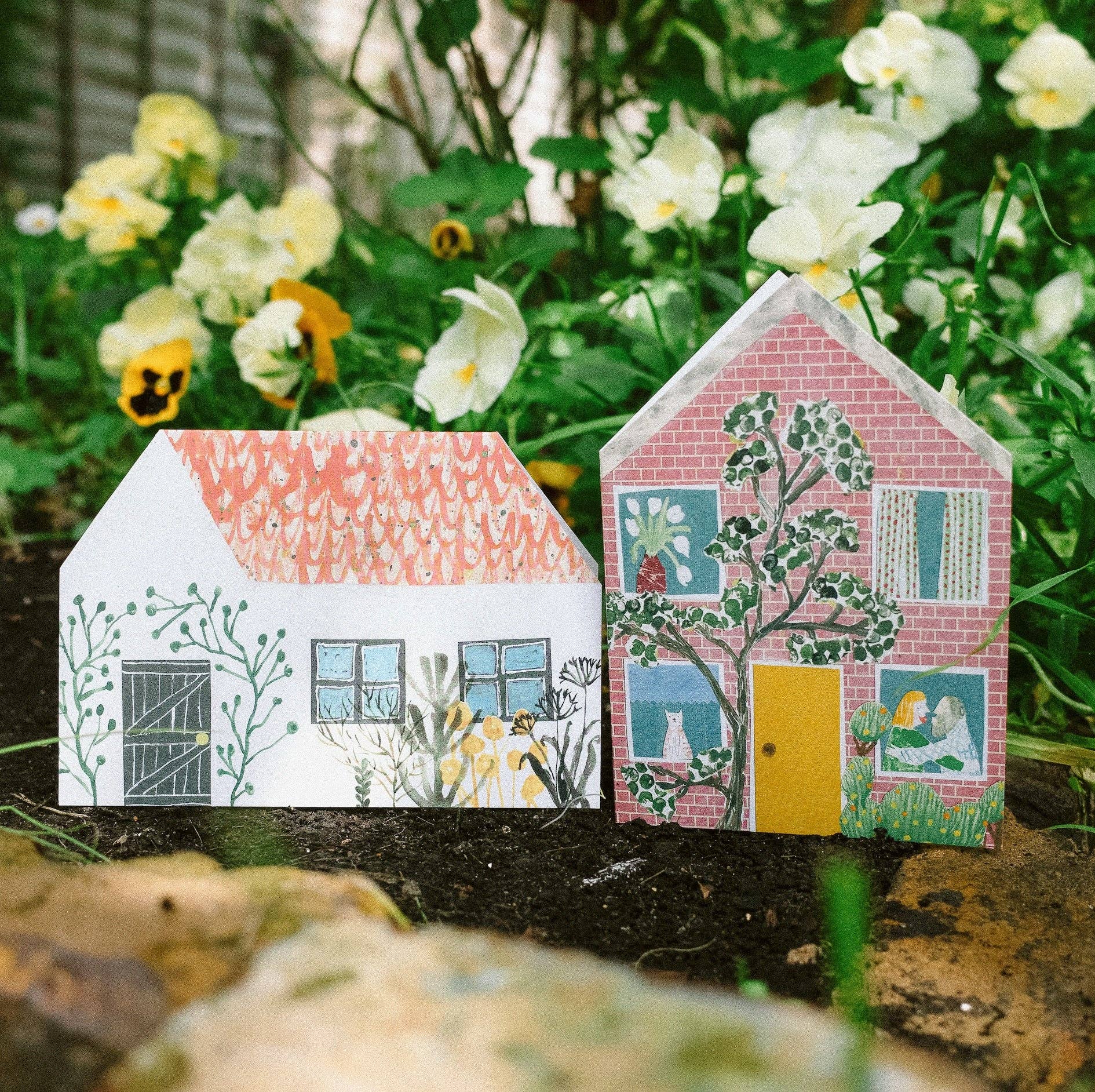 Two greeting card houses with colourful designs on a natural background with flowers.
