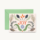 A greeting card with botanical illustrations, shown with a green envelope behind it on a light background. The card features the word 'JOY' in red letters with green leaves, red-orange berries, and a blue-and-orange sun motif.