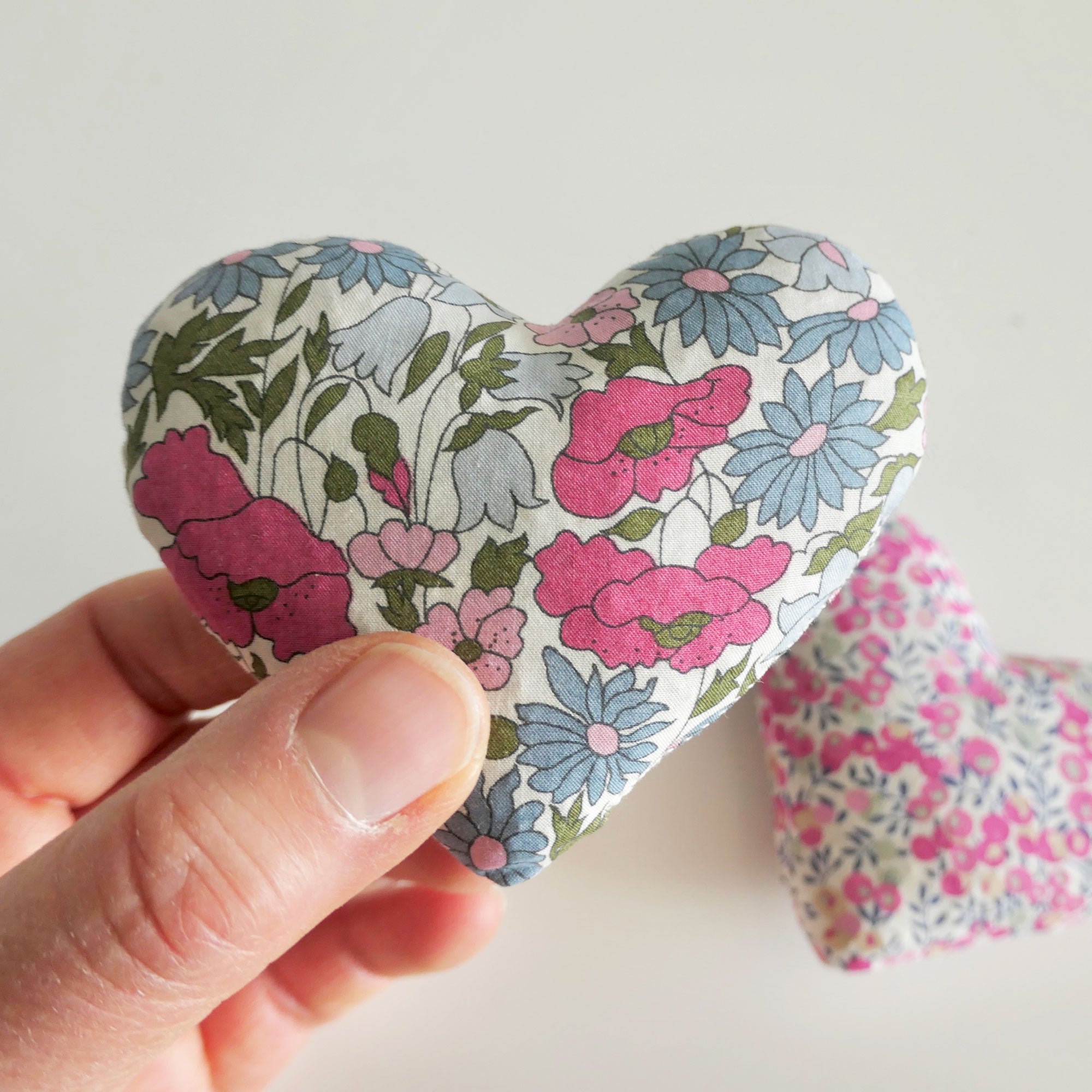 Floral-patterned heart-shaped fabric sachet held by a hand on a light background