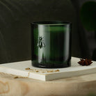 Dark green candle with a bee logo on a wooden surface and book
