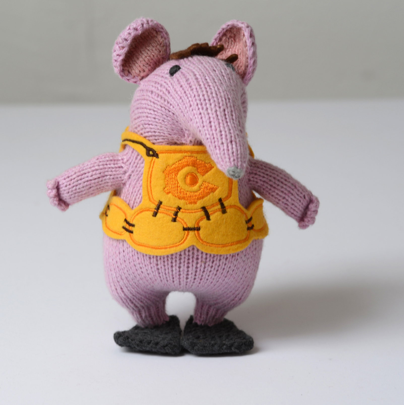 A knitted mouse toy wearing a yellow harness vest and black shoes.