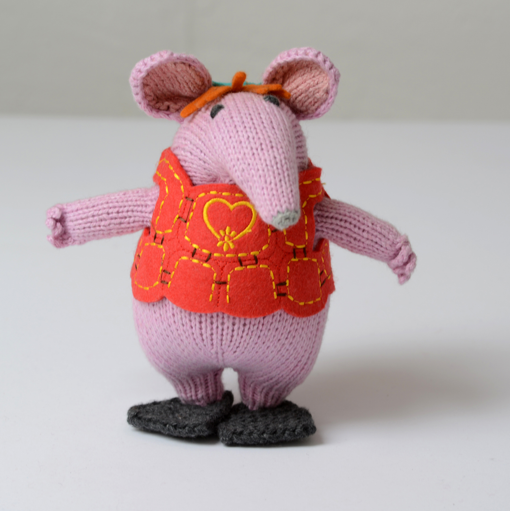 A knitted mouse doll with pink ears and legs, wearing a bright orange-red vest with yellow embroidered details and dark grey shoes, shown centred on a light grey background.