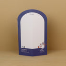 A freestanding arched paper card  with a white writing area and a dark blue border, placed on a light wood surface against a tan background. The bottom section includes printed branding and contact details.