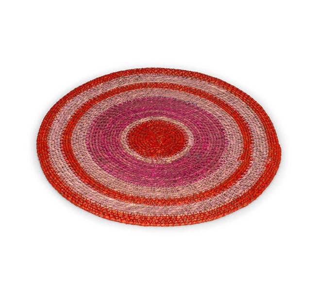 A round woven placemat with concentric rings of red, pink, and beige raffia on a white background.