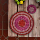 A round woven placemat with concentric rings of red, pink, and beige sits on a rustic wooden table, with a matching small round coaster above it and limes in the corner.